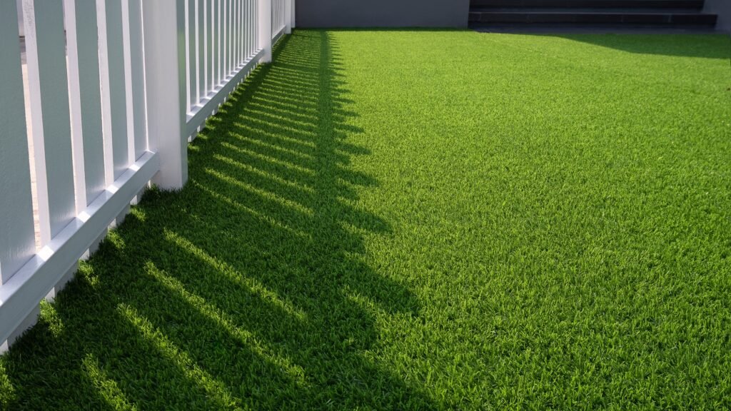 Sunlight and shadow of white wooden fence on green artificial turf surface in front yard of home